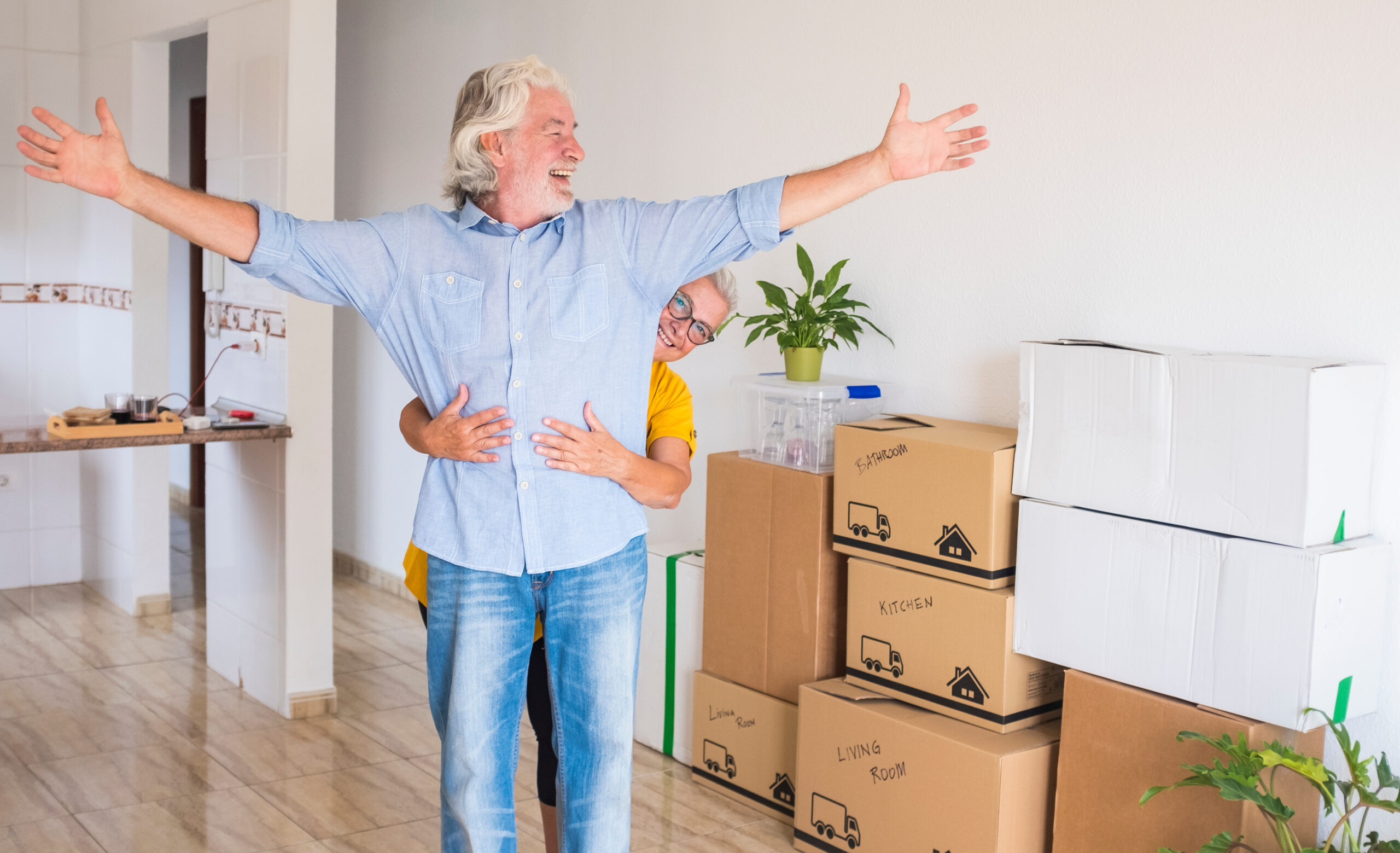 Two cheerful smiling senior people with white hair embraced during relocation, happy for the new beginning like retired with moving boxes on the floor